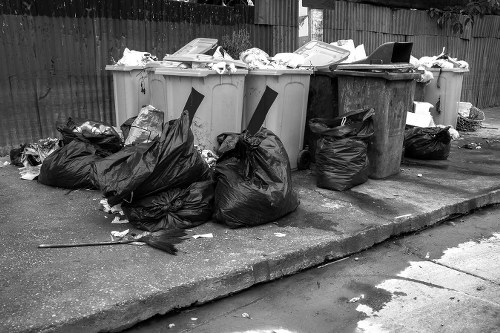 A row of five large wheelie bins positioned on a concrete pavement outside a property, with three of the bins being light grey and two being darker grey or black. The bins are filled with various types of rubbish, with some overflowing and items such as papers and plastic containers visible at the top. Surrounding the bins are multiple tightly packed black plastic rubbish bags, some with their tops gathered and tied, and others partially torn open with loose waste spilling out. The scene is set outdoors on a paved area with a wooden fence in the background, typical of a residential or commercial waste collection point in Balham. The environment appears clean but cluttered due to the accumulated waste awaiting collection, emphasizing the importance of professional rubbish removal services in the area. The overall atmosphere suggests ongoing waste management activities, with the focus on the collection and disposal of mixed household or commercial refuse as managed by Commercial Waste Balham.