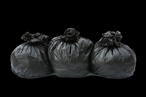 Three large black rubbish bags, made of thick plastic material with a slightly crinkled surface, are positioned side by side on a dark, featureless background. The bags appear full and are tied at the top with twisted closures. The leftmost bag shows some slight bulging, indicating it contains substantial waste, while the middle and right bags are similarly filled and neatly tied. The bags are placed on what looks like a pavement or driveway surface, possibly outside a residential or commercial property in Balham, London. The lighting is even but subdued, highlighting the dark color and textured finish of the plastic bags without casting harsh shadows. This scene is typical of rubbish removal activities in the area, with Commercial Waste Balham often handling collection and disposal of such waste bags for local clients. The environment appears clean and organized, with no visible debris apart from the rubbish bags themselves, emphasizing a straightforward waste disposal process common to many properties in the postcode region.