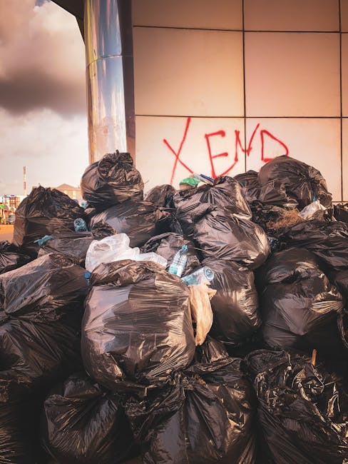 A large accumulation of black rubbish bags filled with waste is stacked against a modern building wall with light-colored tiles. Some bags appear partially torn, revealing discarded plastic bottles an