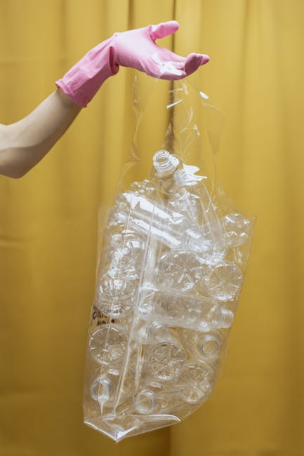 A large accumulation of black rubbish bags filled with waste is stacked against a modern building wall with light-colored tiles. Some bags appear partially torn, revealing discarded plastic bottles an