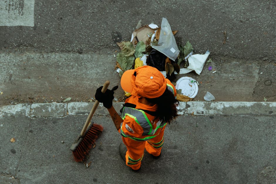 A worker dressed in high-visibility orange safety clothing and a matching orange hat is standing on a concrete pavement, facing a low concrete wall. The worker is holding a large broom with a long woo