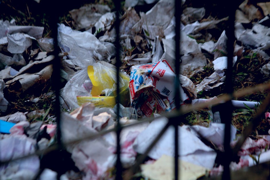 A close-up view of a small section of a black metal fence with vertical bars, behind which piles of discarded rubbish are visible on the ground. The rubbish includes crumpled paper, plastic bags, and