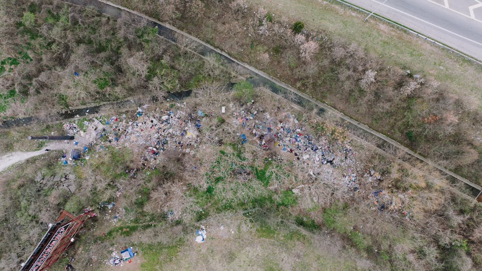 A close-up view of a small section of a black metal fence with vertical bars, behind which piles of discarded rubbish are visible on the ground. The rubbish includes crumpled paper, plastic bags, and